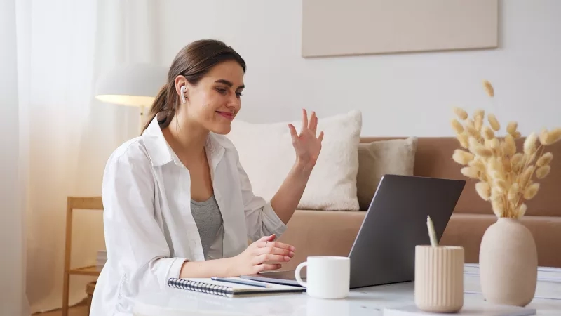 Woman in a white shirt having a video call on her laptop in a living room.
