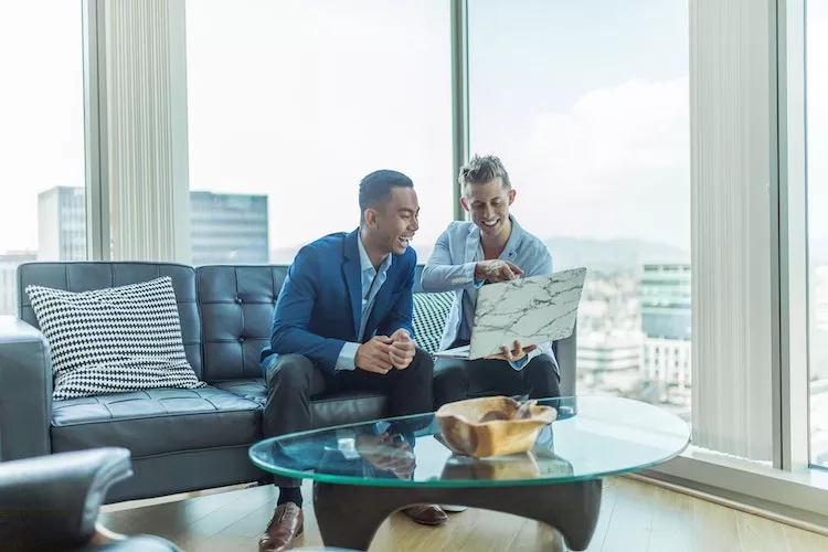 Two men in suits sitting on a couch in a modern room with a view of a city.