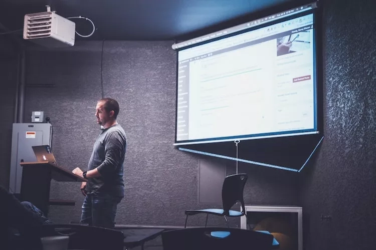 A man stands behind a podium giving a presentation on a large screen in a conference room.