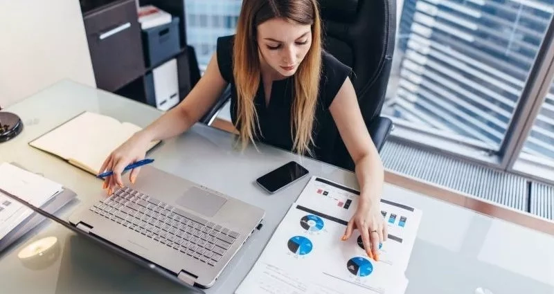 A woman working on a laptop with papers on a glass table in a corporate office.