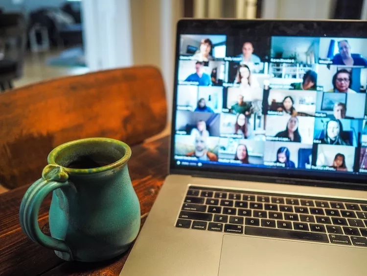 Laptop with video conference on screen and cup of coffee on a table in front of it.