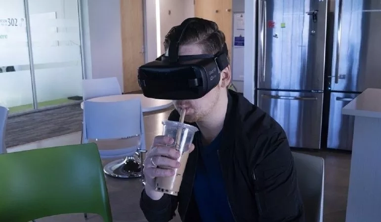 A man in a black jacket and virtual reality glasses sips a latte in a lounge.