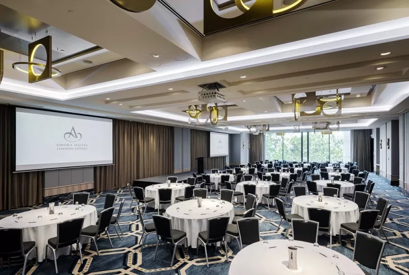 Amore Hotel Jamison Sydney's Whiteley Ballroom, with large screens, a podium, and a carpeted floor with chairs.