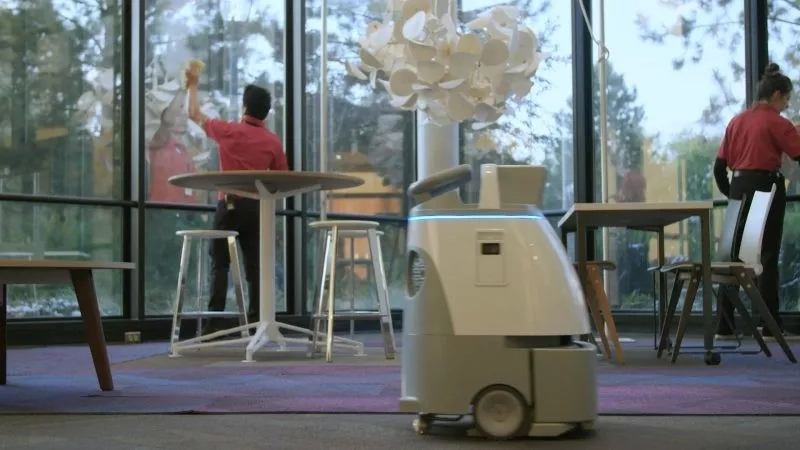 Two people are cleaning glass windows while a robot vacuum cleans the floor.