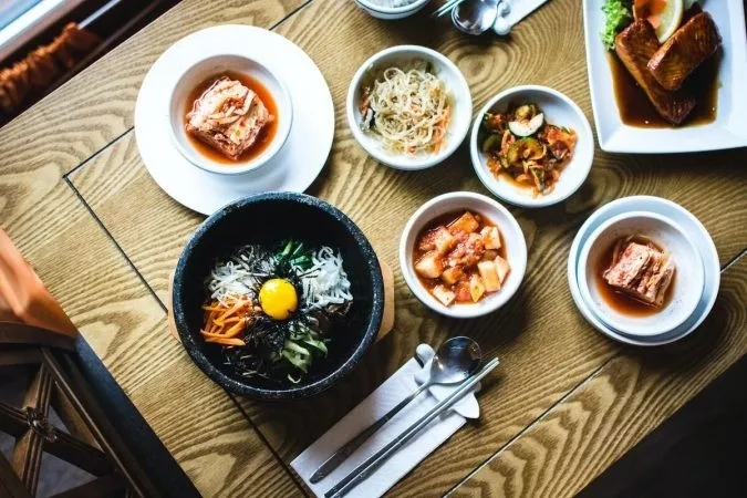 Korean food dishes on a table with a bowl of kimchi and a bowl of noodles with a spoon.