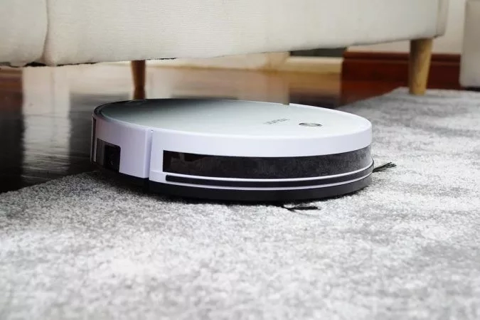 White robotic vacuum cleaner placed on gray carpet under white couch in a room with wooden floor.