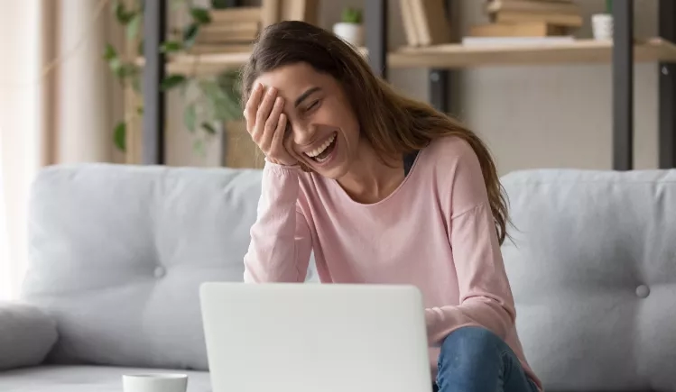 A woman laughs while sitting on a couch with a laptop on her lap.
