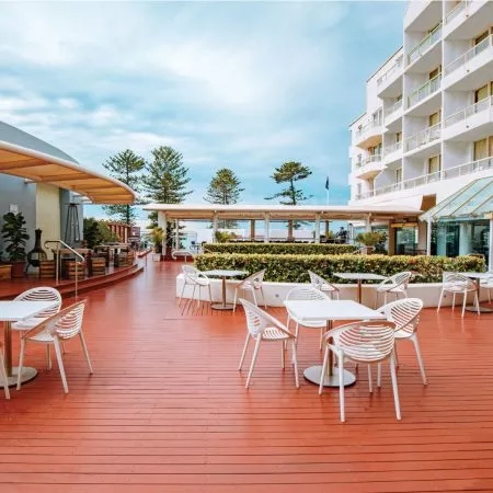 Novotel exterior with wooden deck, white chairs, and tables, surrounded by lush landscaping under a clear sky.