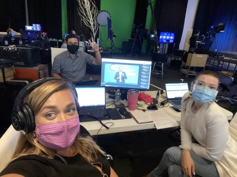 Two women and a man wearing masks are in a studio with multiple monitors and a green screen.