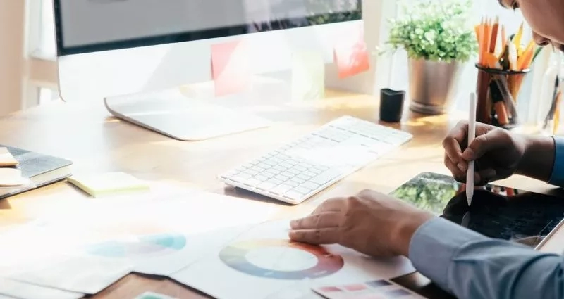 Person writing in notebook with color palette, tablet, and keyboard on desk in front of computer.