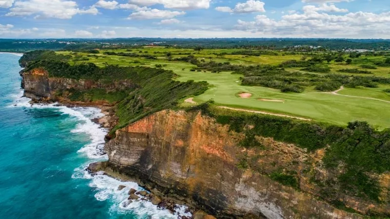 Aerial view of the golf course with a cliff overlooking the ocean, lush greenery, and clear blue sky.