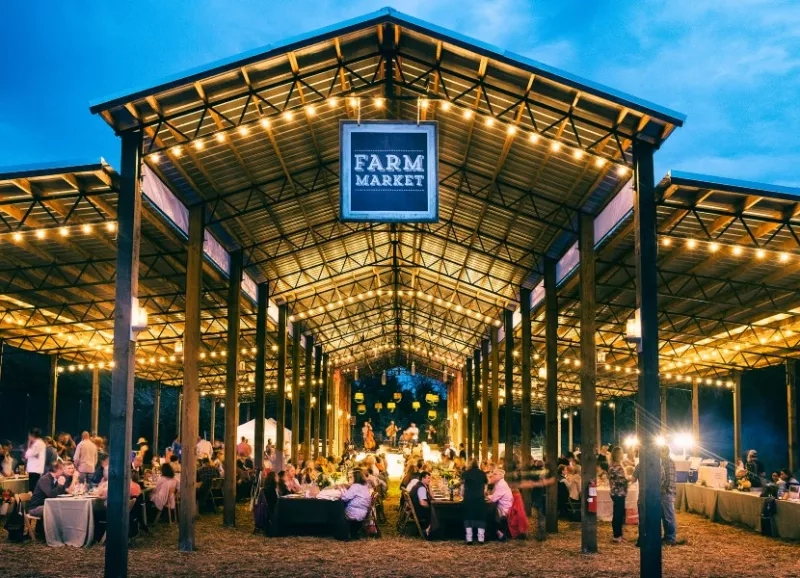 A farm market barn with tables and string lights, people are gathered and a stage is visible.