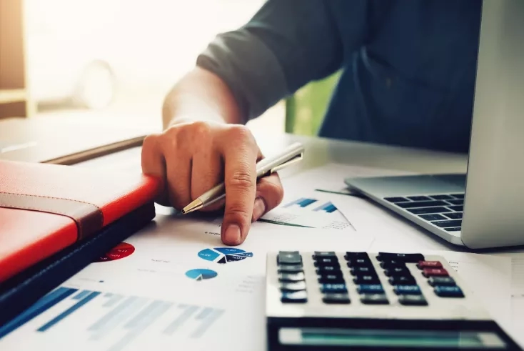 Hand holding a pen over a calculator with a laptop and paper charts on the desk.