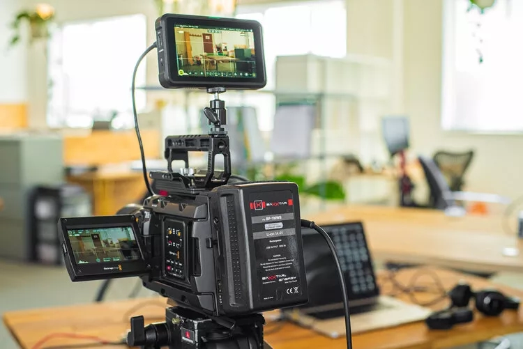 A black video camera with a screen on top is on a tripod in an office.