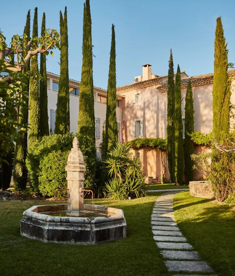 A charming garden with a fountain, a stone path, and a cozy building at Château de Fonscolombe.