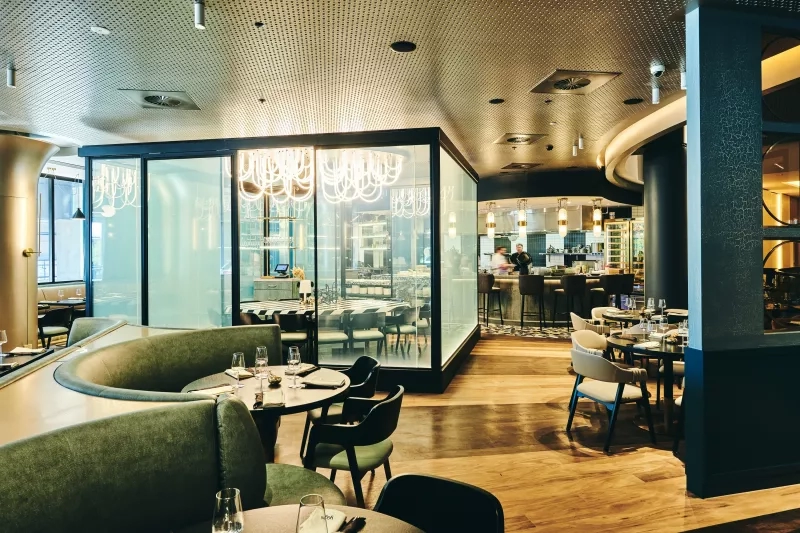 View of the dining area of Yugo, featuring booths, tables, chairs, and a glass-enclosed kitchen with chandeliers.