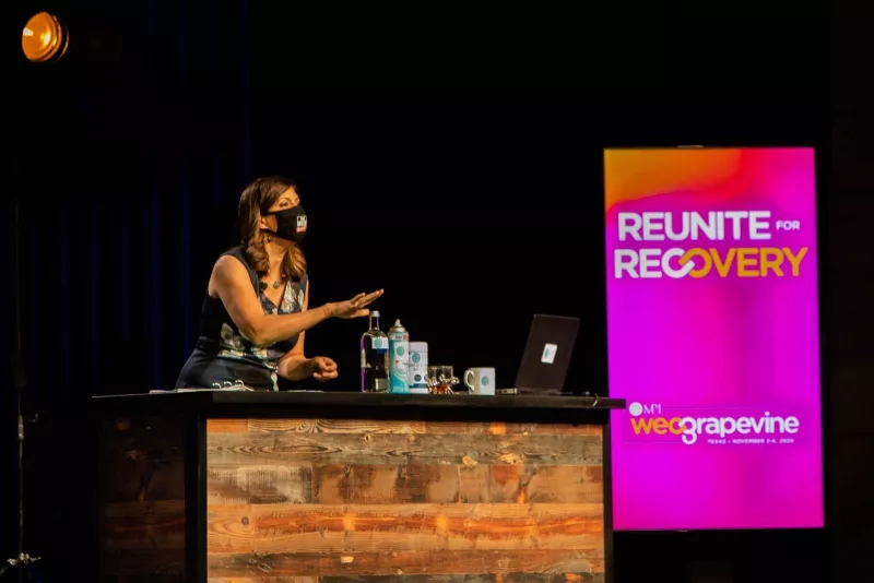 A woman behind a counter giving a speech with a screen that says Reuniting for Recovery