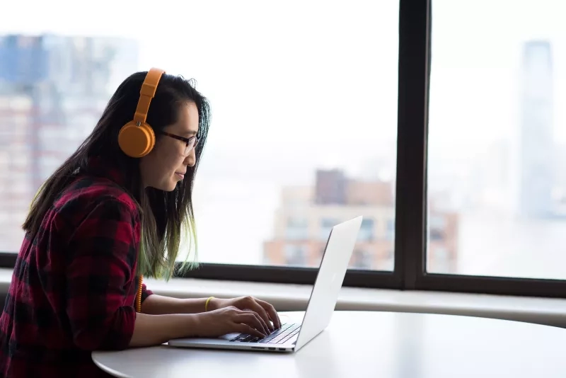Woman wearing headphones and using a laptop in front of a window with a city view.