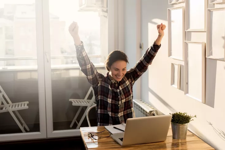 A woman is smiling at a desk with a laptop, pen, and potted plant at her home.
