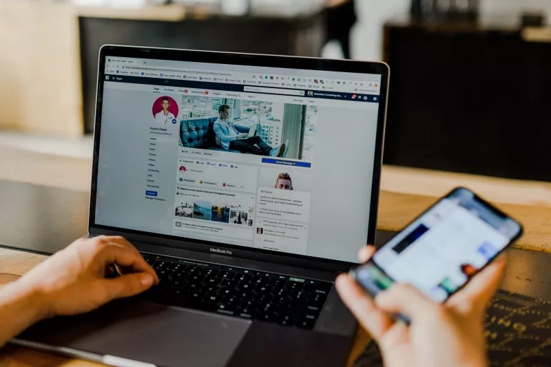 Person using a laptop and mobile phone to check Facebook while sitting in a wooden desk.