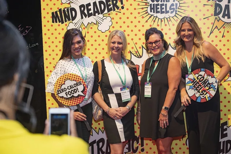 Four women posing in front of a comic book theme backdrop at a conference event.