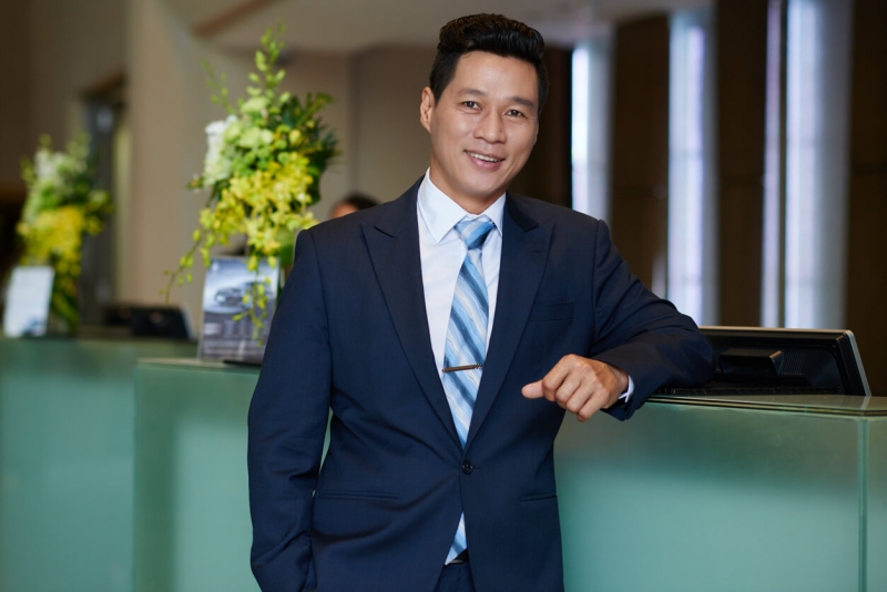 Hotel owner with hand in pocket leaning on front desk and looking at camera