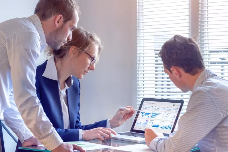 Three people looking at a laptop showing a dashboard, one pointing with a pen.