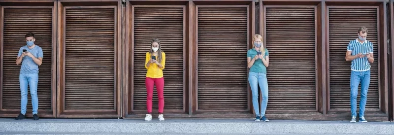 Four people wearing masks stand apart from one another while looking at their phones.