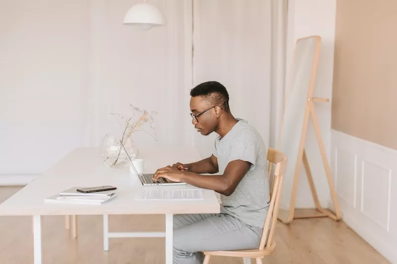 African American man working at home at desk with laptop and books on table, looking at screen.