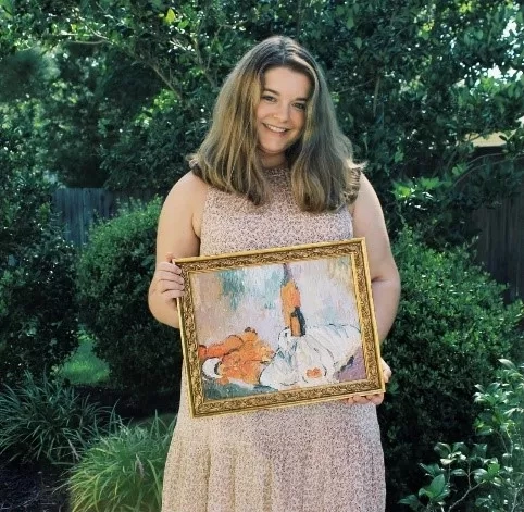 A woman stands outdoors holding an oil painting of a cat with trees and bushes in the background.