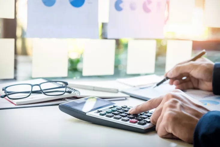 Person using calculator to calculate budget, with papers and glasses on desk in blurred office background.