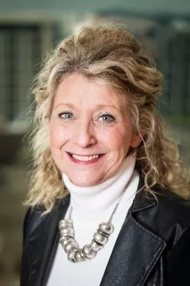 Cindi, a woman with blonde hair and a black jacket with a necklace, is smiling for a photo.