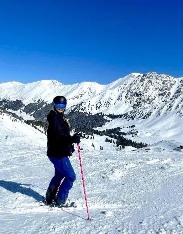 A girl with helmet and goggles standing on skis with snowy mountains in the background.