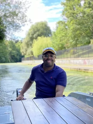 Keegan wearing a blue cap and shirt sitting in a boat on a river in London.