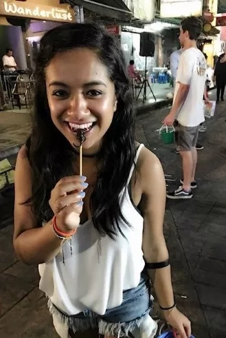 A woman in a white shirt and denim shorts smiles while eating at Wanderlust in Thailand.