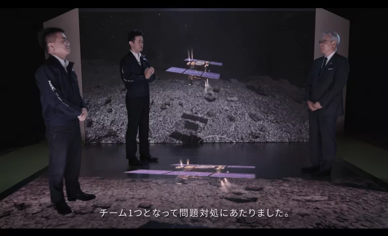 Three men are standing in front of a projection of a satellite on the moon's surface.