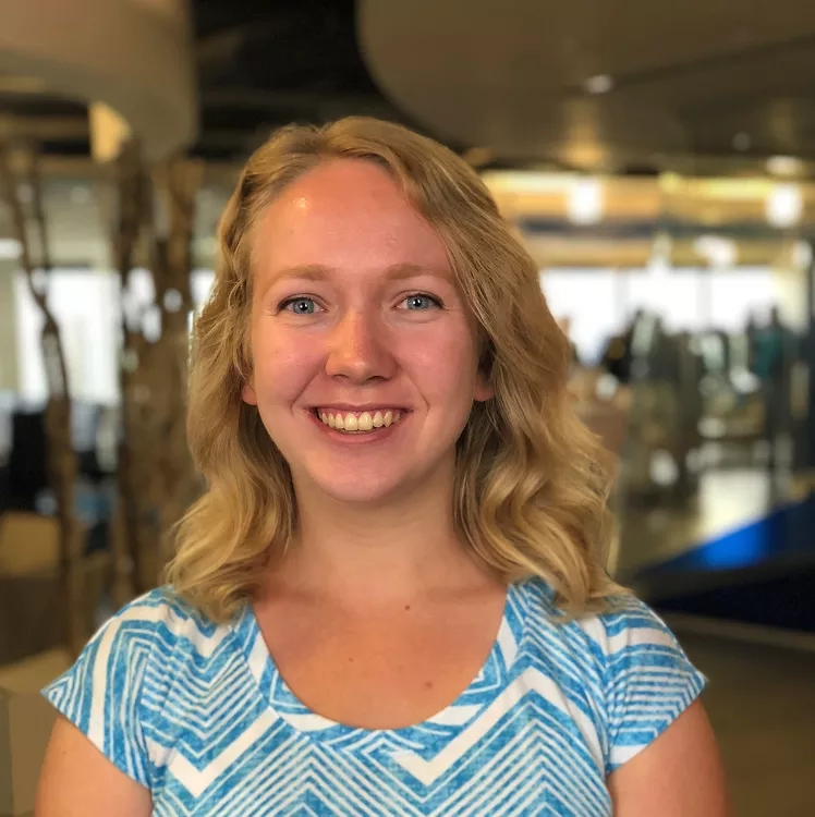 A smiling woman in a blue shirt stands in an indoor space with blurred background, possibly an office.