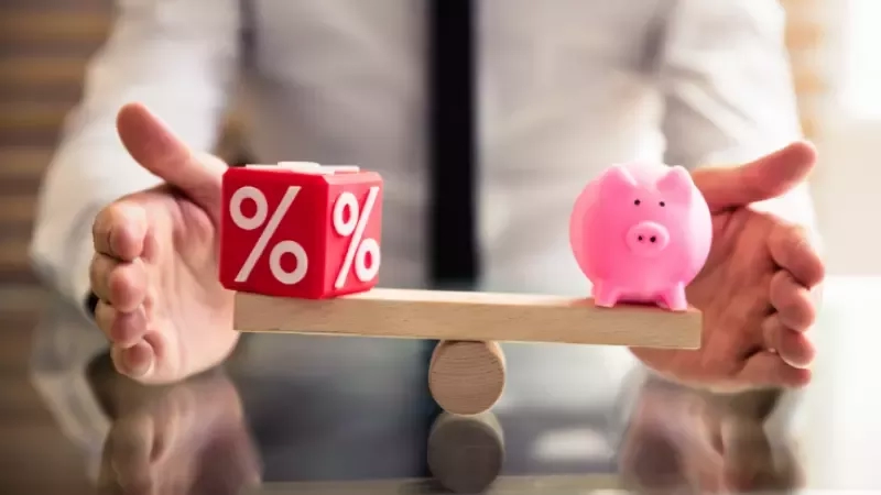 A piggy bank and a percentage symbol on a seesaw, held by a man in an office.