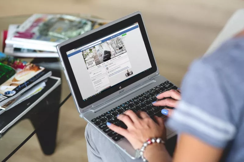 A woman is typing on a laptop with blue nails while sitting on a couch next to a coffee table.