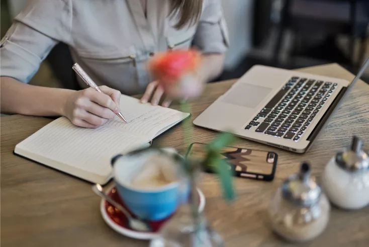 Woman working at a table with a laptop, pen and paper and a coffee.