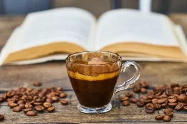 A cup of coffee with coffee beans and an open book on a wooden surface.