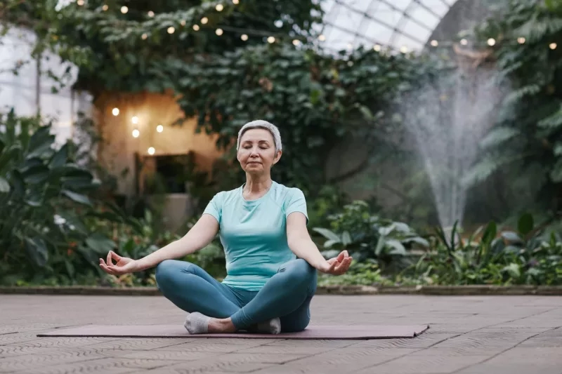 A woman meditating in a lotus position on a yoga mat in a lush, indoor tropical garden.