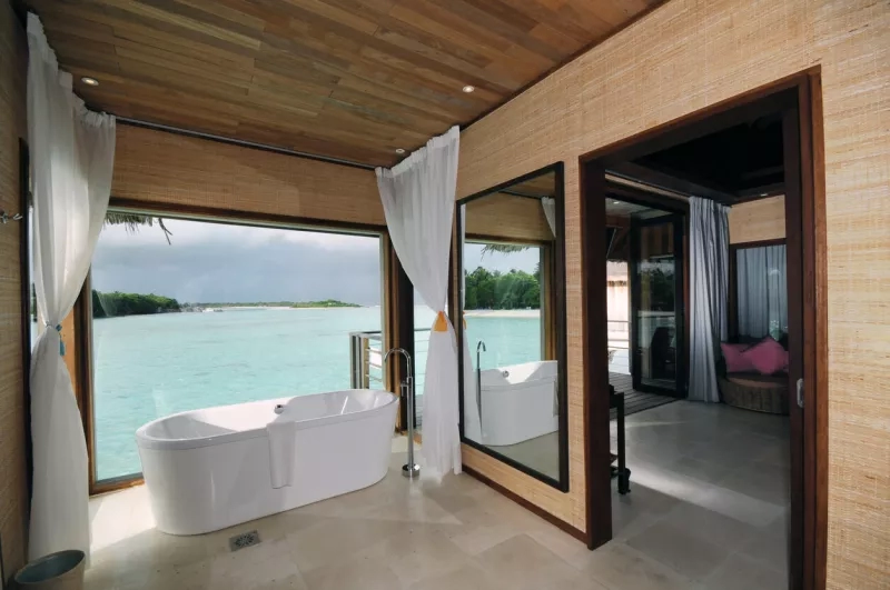 A modern bathroom with a bathtub overlooking the ocean, tiled floor, and wooden ceiling.