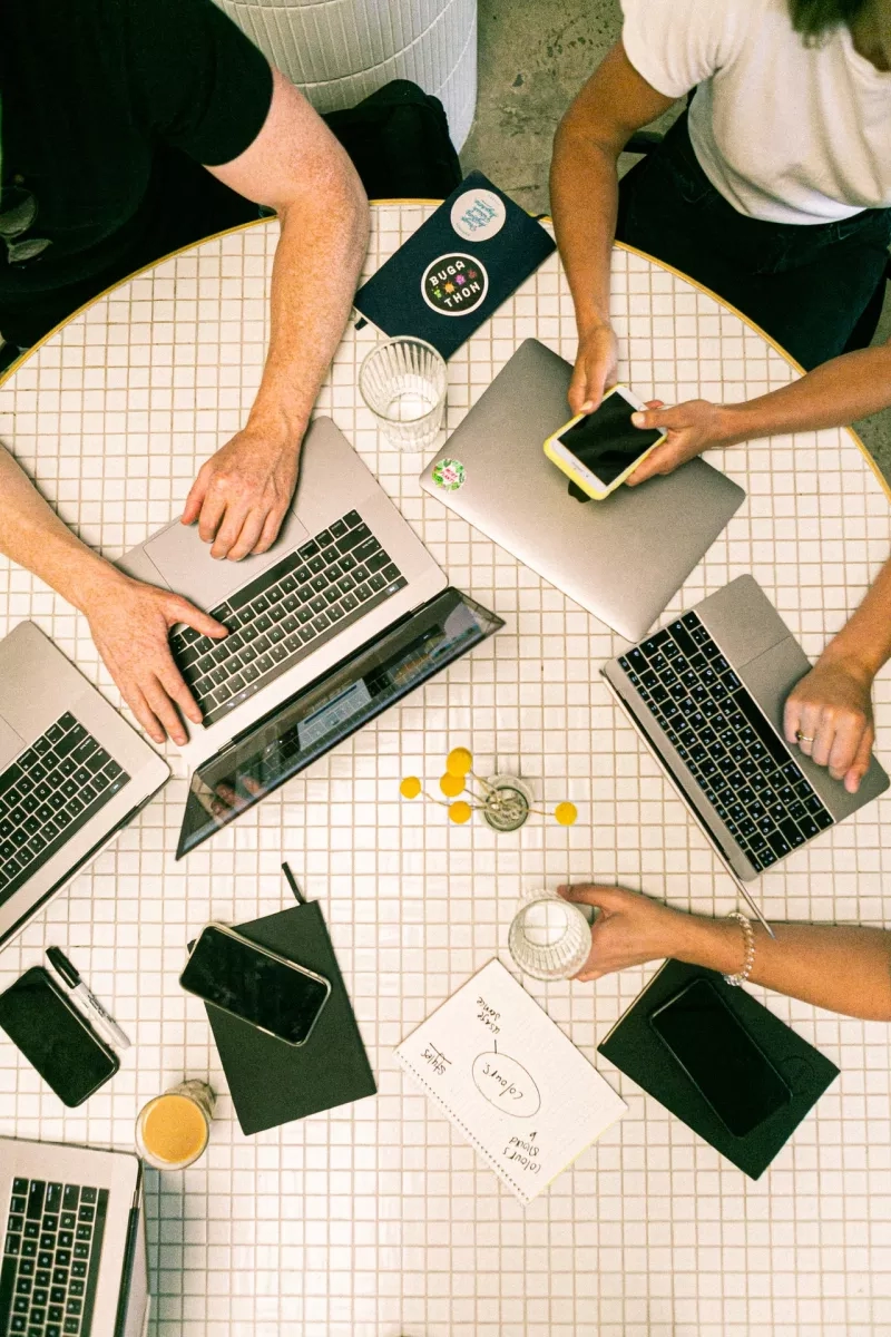 Five people sit around a table with laptops and papers, engaged in work or a meeting.