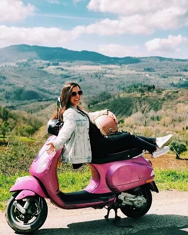 A woman on a pink Vespa scooter with mountains in the background.