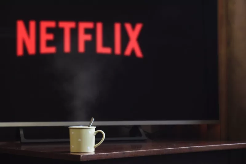 A Netflix logo on a TV screen with a steaming cup of coffee on a desk in front of it.