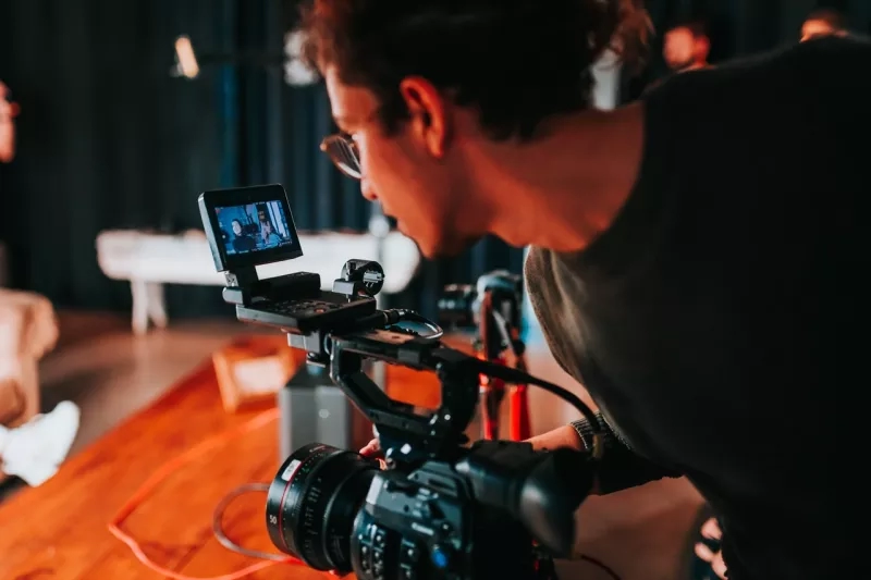 Person with glasses and dark shirt operating camera with small screen and cord on wooden table.