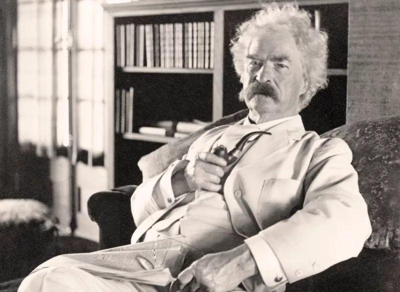A photo of Mark Twain with a pipe in his hand, sitting in a chair in his study.