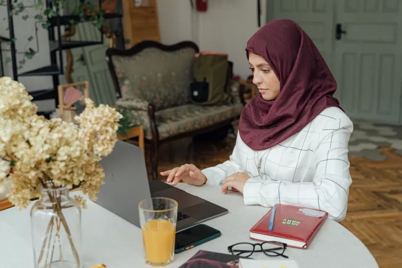 A woman wearing hijab is working on a laptop on a table with glasses, book, and glass of orange juice.