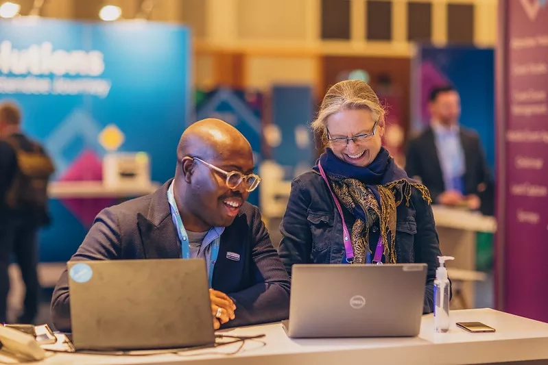 Two people are sitting at a table, laughing while working on their laptops at a Solutions Conference.